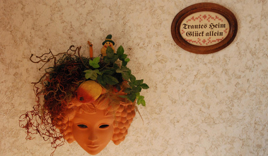 Theatermaske, orange, aufgehängt an Wand, oben rechts darüber ein gesticktes, ovales Schild Trautes heim, Glück allein